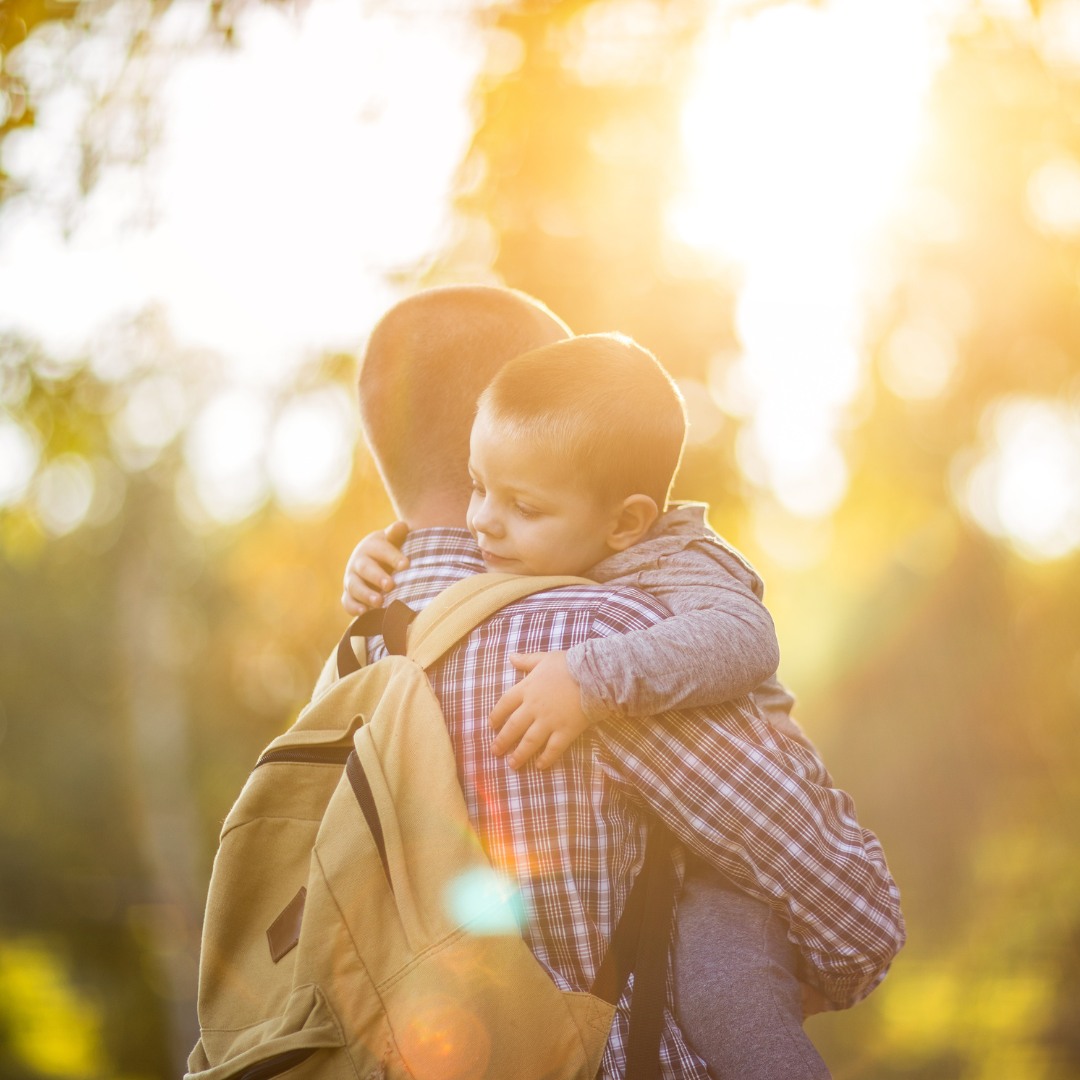 The father embraces his young son, holding him up for a hug as filtered sunlight filters through trees